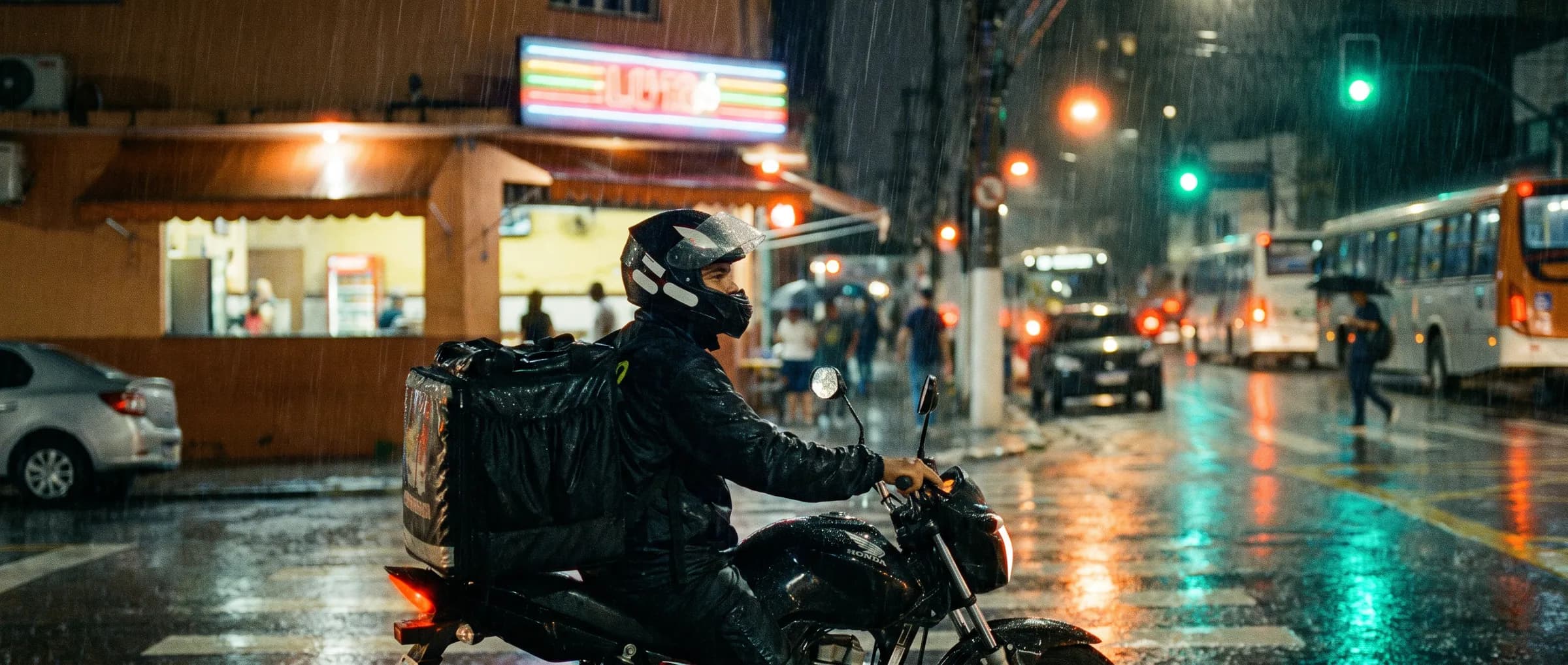 Motoboy de entrega parado na chuva, à noite, em frente a uma lanchonete